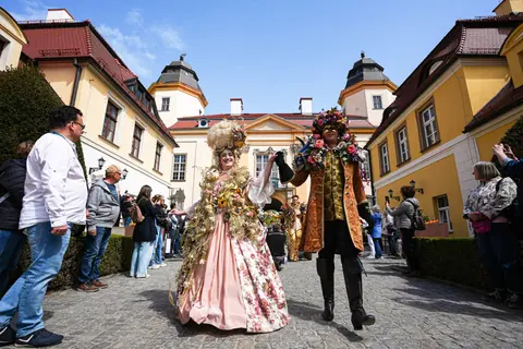 Postaci w kwiatowych kreacjach z epoki spacerują między zamkowymi budynkami