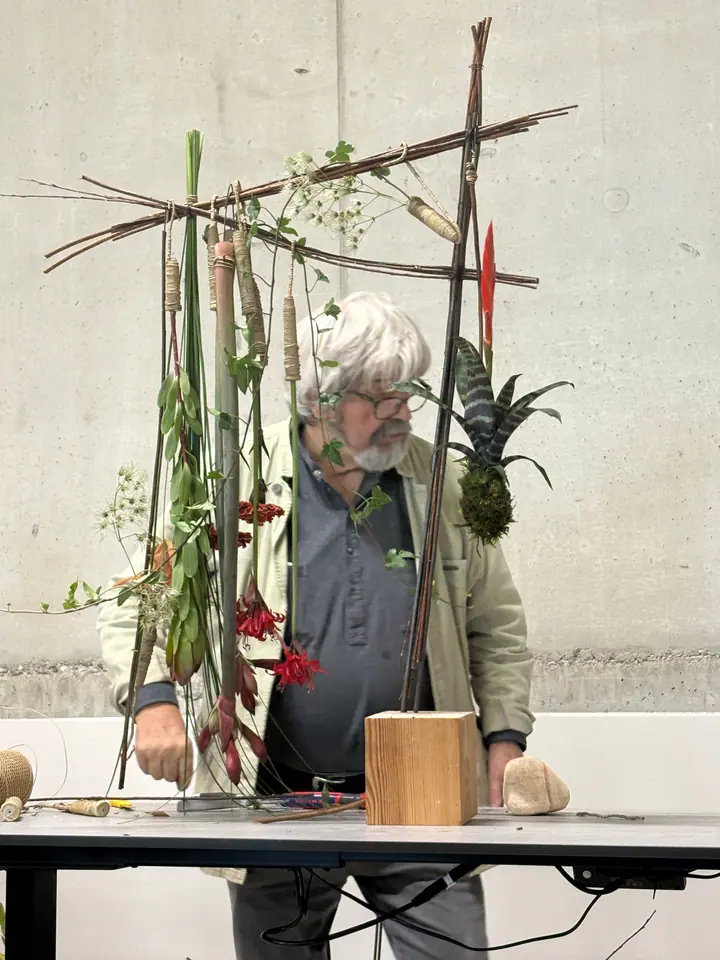Gregor Lersch za konstrukcją z gałęzi, z której zwisają owinięte sznurkiem łodygi, kwiaty i bromelia w kokedamie